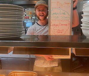 A young man smiling broadly, standing behind a serving counter offering fried seafood. He is holding up a menu board that reads: "Uncle Zack's Fish Frenzy: Garlic/Lime Prawn Skewers, Lemon/Cracked P. Squid Game"