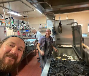 A woman wearing a striped apron, smiling at the camera as she fries eggs on a large industrial hotplate. A bearded man 'photobombs' the picture, his smiling face in the corner foreground