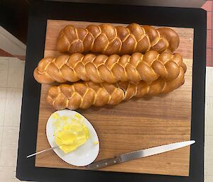Three long, artfully braided loaves of bread lying on a wooden serving board, alongside a plate of butter and a bread knife