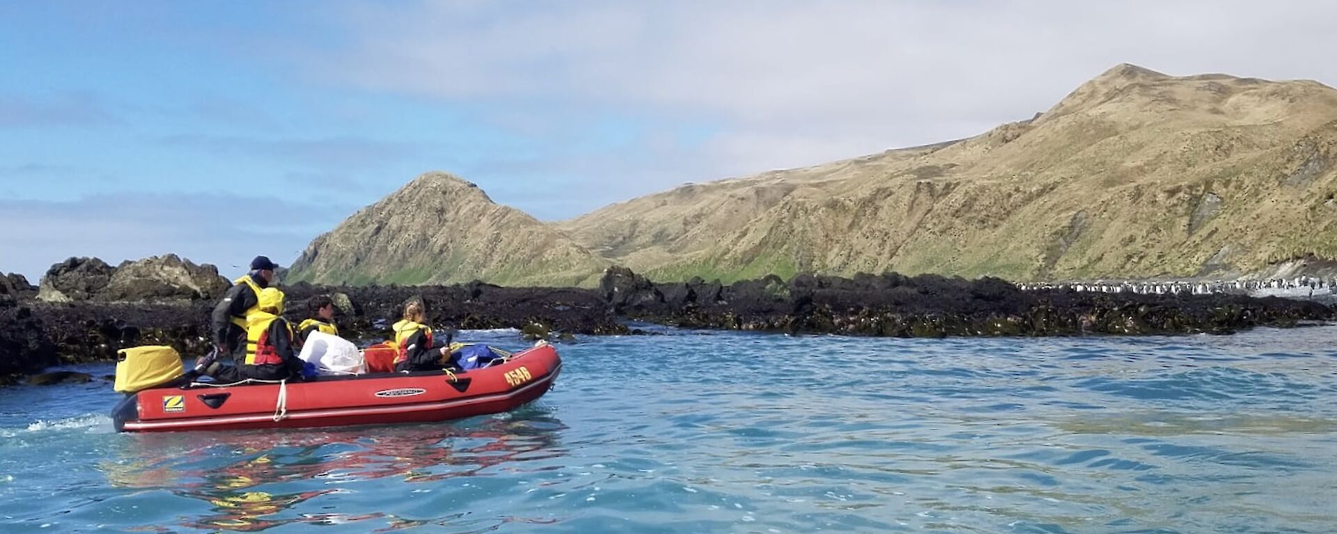 A group of people in a red inflatable boat cruise past the shore of a rocky island