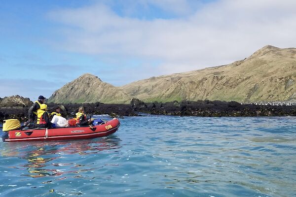 A group of people in a red inflatable boat cruise past the shore of a rocky island