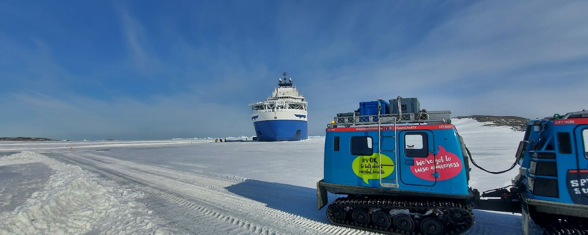 A blue Hägglunds vehicle drives across the sea ice out to a blue and white ship