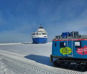A blue Hägglunds vehicle drives across the sea ice out to a blue and white ship