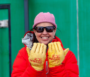 A woman in a red jacket in front of a green building holding her hands up in bright yellow gloves