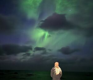 A lady stands in front a green aurora in the cloudy sky