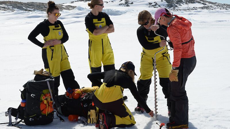 Four people dressed in yellow survival suits follow the instructions of a woman in red to use a sea ice drill