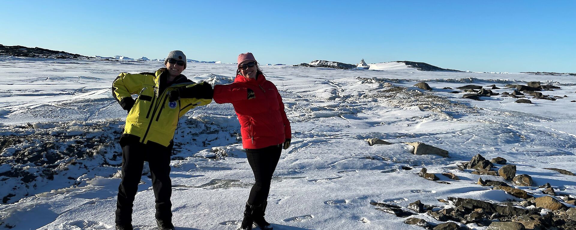 Two smiling women with elbows touching. Sea-ice and icebergs in the background