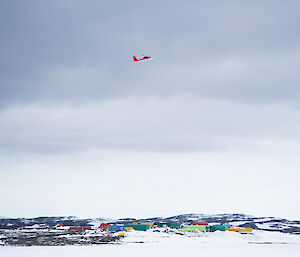 A small red plane flying above buildings in a snowy landscape