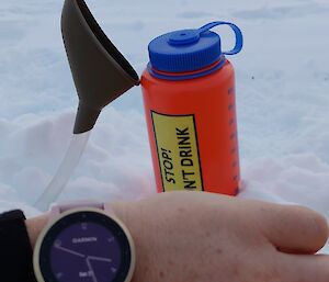 A bare hand with wristwatch in foreground and drink bottle in snow in background