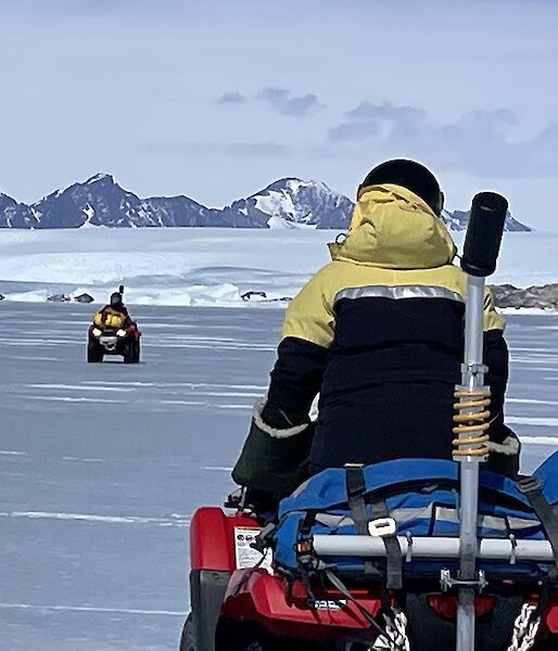 Two people on red quad bikes drive across the sea ice