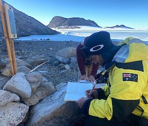 A person in a yellow jacket writes in a visitor type book on a rocky shoreline