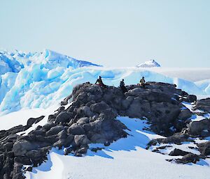 Three people relax in the sun on top of a rocky outcrop with ice all around