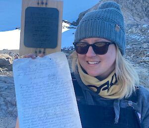 A women in sunglasses and beanie holds up a piece of paper with writing on it