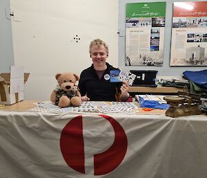 Teddy Bear sitting on a table next to a  smilingman seated in a chair holding some stamps