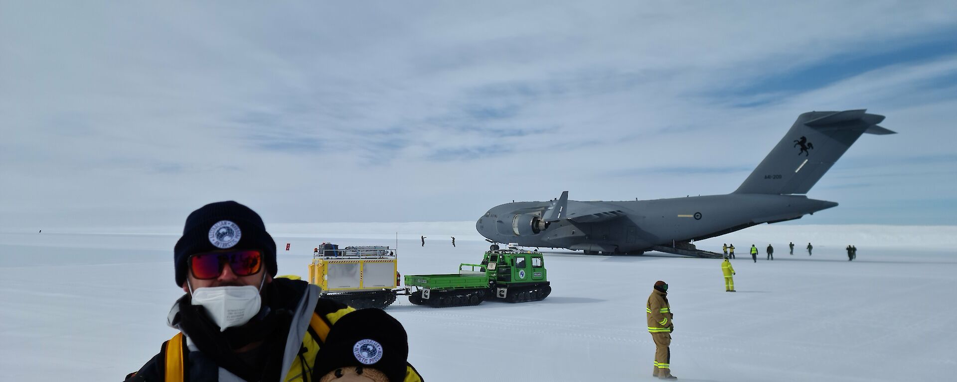 Man in yellow jacket holds teddy bear while standing in front of a very large grey aeroplane