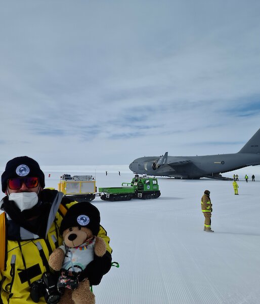 Man in yellow jacket holds teddy bear while standing in front of a very large grey aeroplane