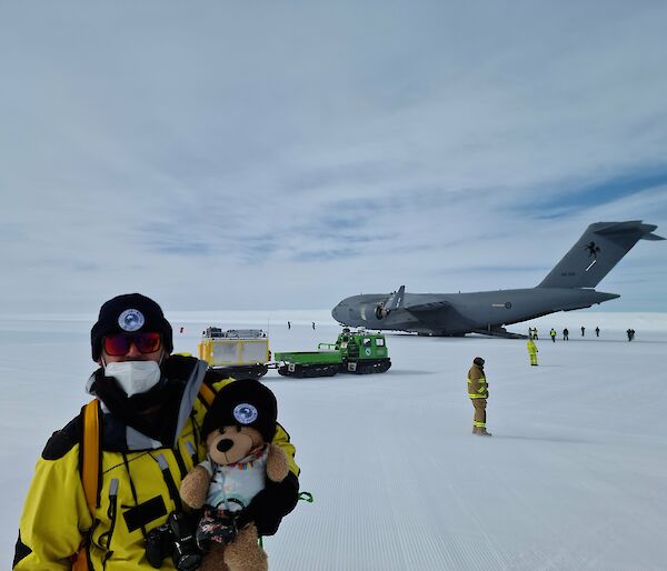 Man in yellow jacket holds teddy bear while standing in front of a very large grey aeroplane