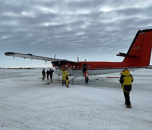 A red and white twin propeller aircraft on the sea ice with seven people standing around ready to board