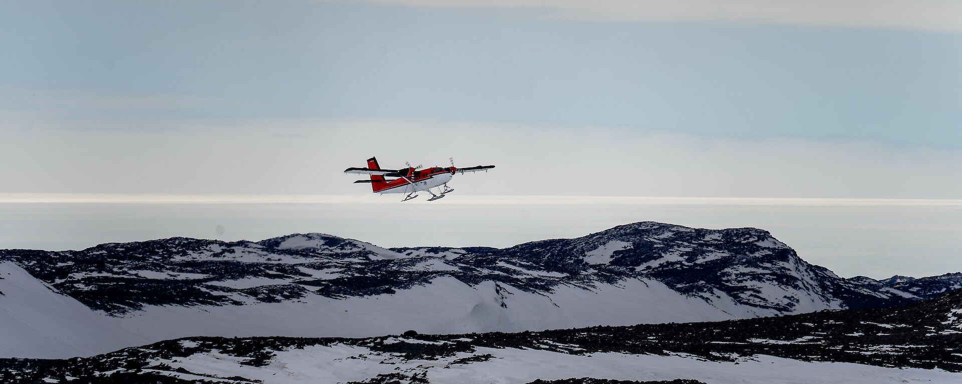 A ski-mounted red and white twin propeller plane in the air just after take-off above an ice-runway with a yellow windsock in the foreground and snowy hills in the background.