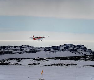 A ski-mounted red and white twin propeller plane in the air just after take-off above an ice-runway with a yellow windsock in the foreground and snowy hills in the background.