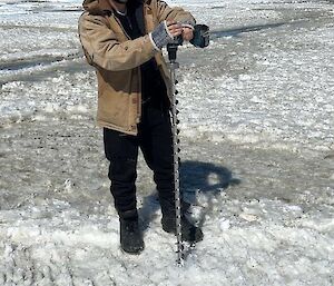 A man in a thick brown jacket and beanie uses a drill with a long drill bit over half his height to drill a hole in the sea-ice on which he stands