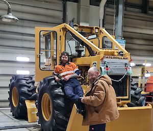 Two men in workshop, one leaning on wheel of digger and one standing in front, both smiling to camera