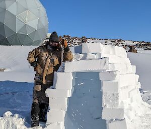 Man stands leaning on igloo made of cut blocks of compressed snow