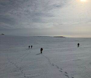 Some individuals walking across the snow with the sun overhead shining through high clouds