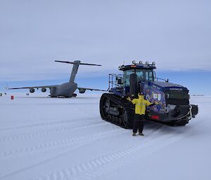 A person in a yellow jacket stands next to a tractor on an ice runway, with a large grey plane behind