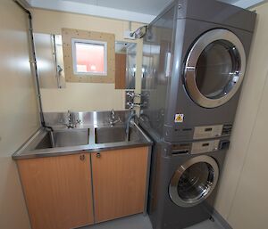 A small laundry room, with stacked washing machine and dryer next to a cupboard with two small stainless steel sinks.