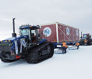 A tractor pulls a red container on sleds. People in hi-vis vests stand next to the container.