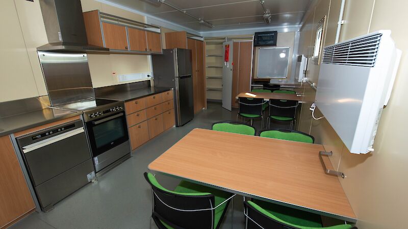 View of the kitchen in the traverse dining/living van, showing stainless steel appliances, cupboards and two dining tables with black and green chairs.