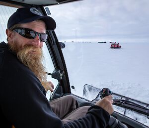 An expeditioner wearing a cap and sunglasses smiles for the camera sitting in the driver's cab of a snow groomer.