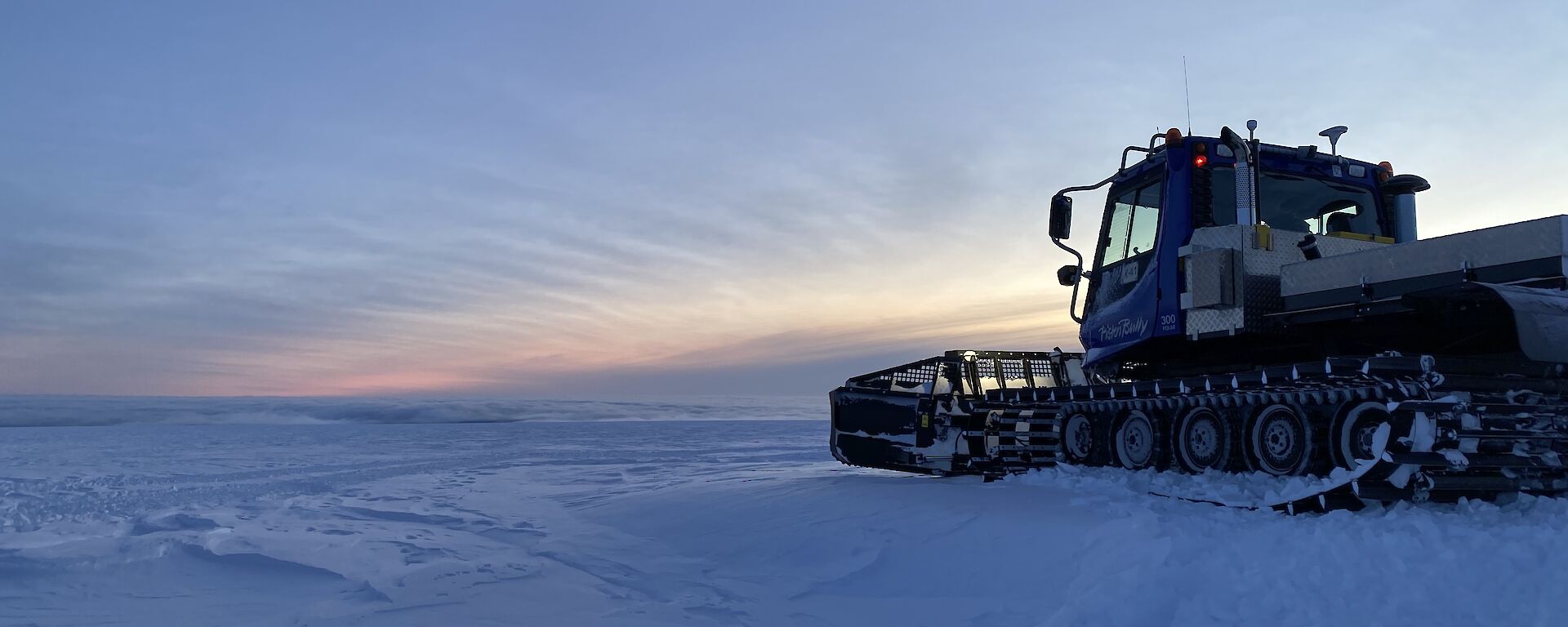 A snow groomer vehicle smoothing out an area on a wide, snowy plain under a twilight sky.