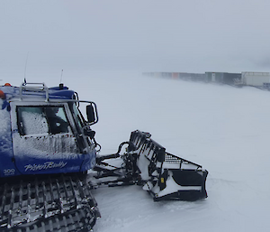 A blue shoveled vehicle sits alongside a row of containers on sleds, amongst thick snow