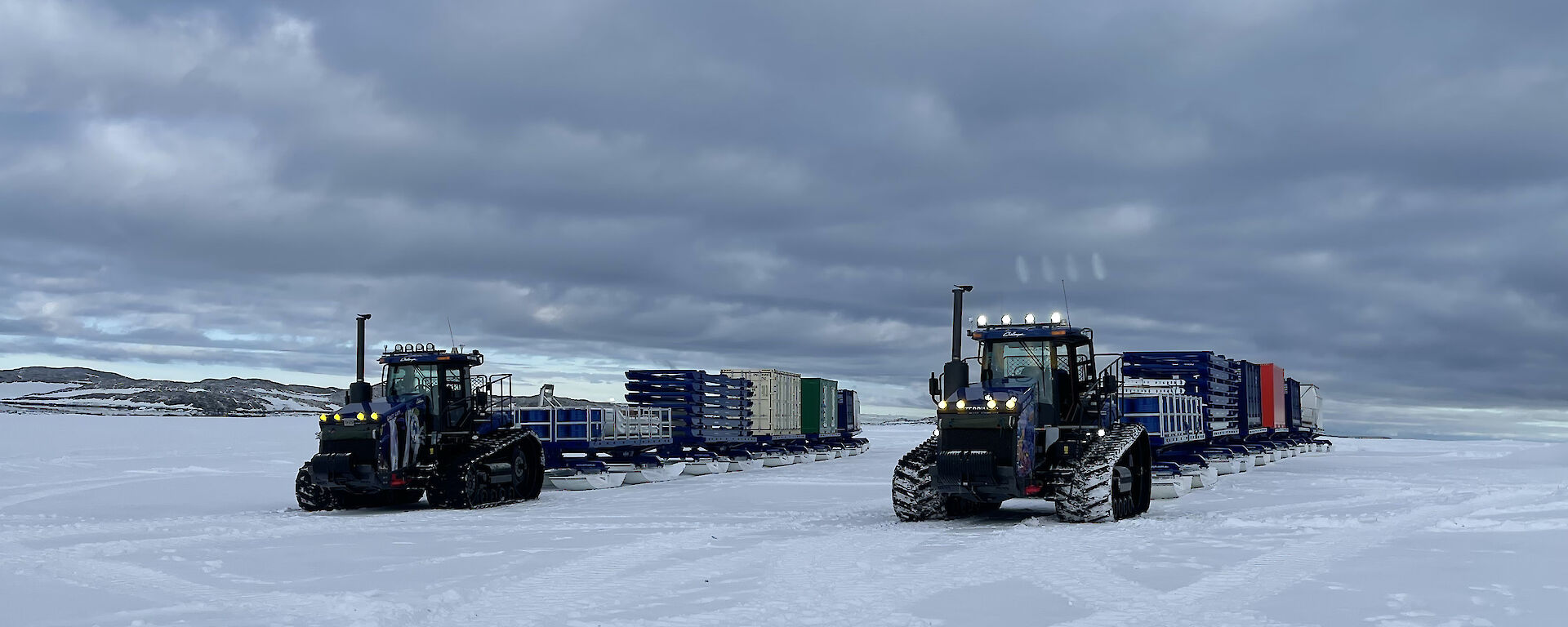 2 tractors tow long trains of shipping containers on sleds across the ice.
