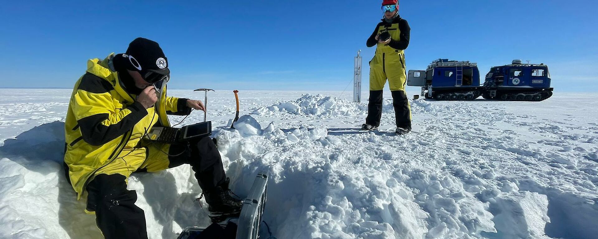 Scientists Tobias Stål and Anya Reading checking data on a field laptop buried in the snow. Blue Hagglunds vehicle in the background