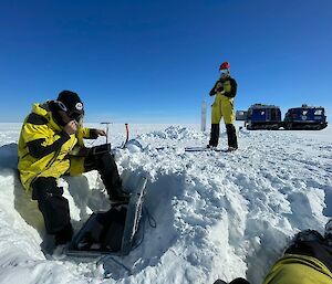 Scientists Tobias Stål and Anya Reading checking data on a field laptop buried in the snow. Blue Hagglunds vehicle in the background