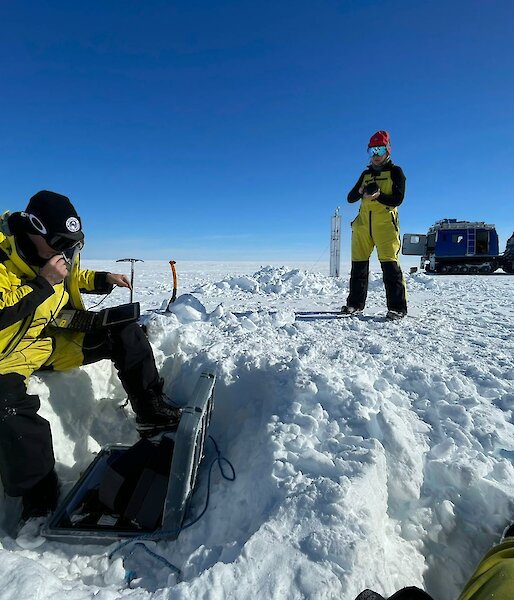 Scientists Tobias Stål and Anya Reading checking data on a field laptop buried in the snow. Blue Hagglunds vehicle in the background