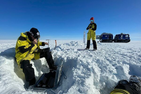 Scientists Tobias Stål and Anya Reading checking data on a field laptop buried in the snow. Blue Hagglunds vehicle in the background