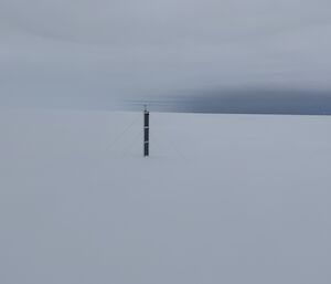 A black, long solar panel attached to a seismometer points upwards in solitary against a white, snowy landscape and white, cloud-filled sky.