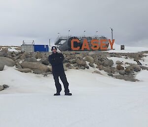 A man standing on snow with crossed arms, looking cold with the Casey station sign in the background