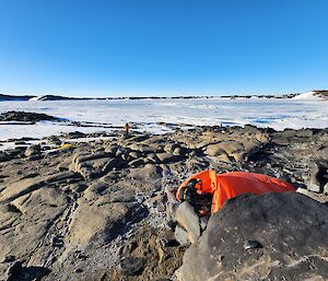 A large orange nylon bag with a sleeping bag inside, on the ground amongst rocks, with snow covered sea-ice in the background