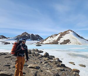 A man wearing a backpack and warm winter clothes stands on rocks with snow covered hills in the background