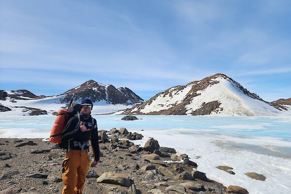 A man wearing a backpack and warm winter clothes stands on rocks with snow covered hills in the background