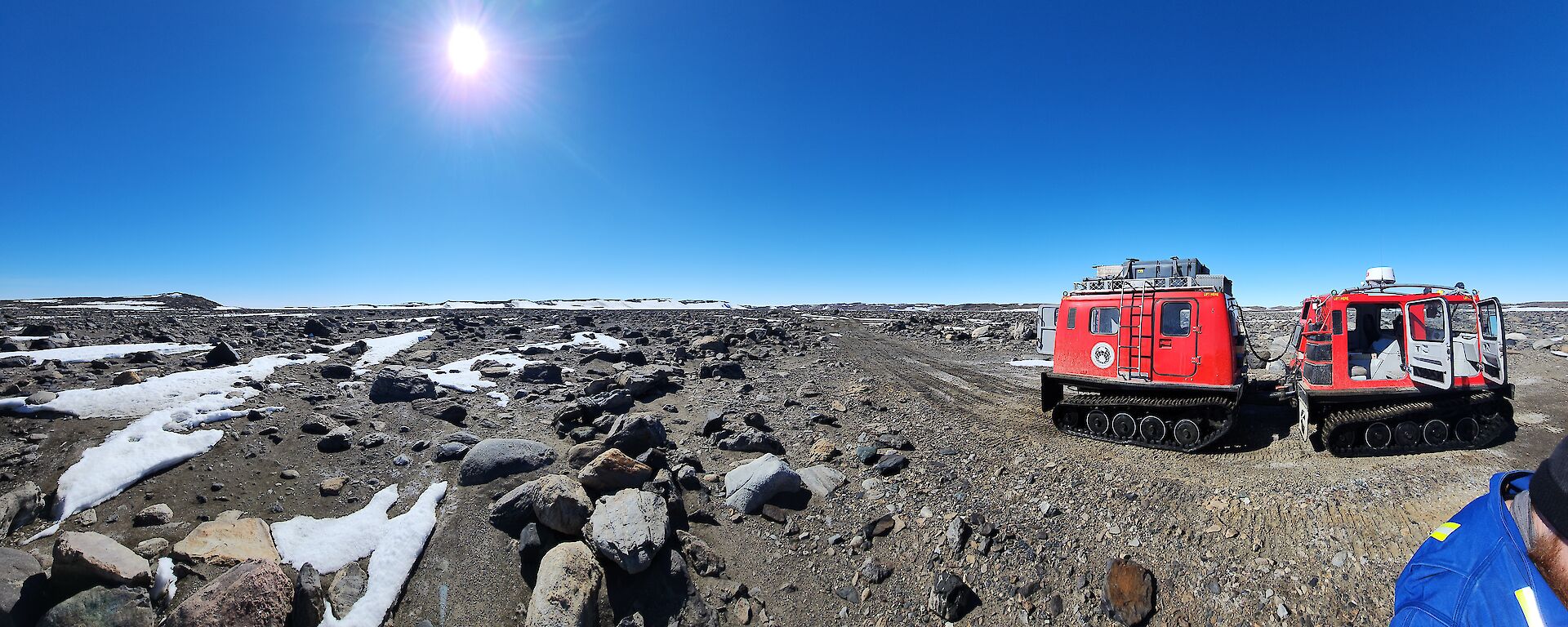 A red box-shaped vehicle towing a red box-shaped trailer, both on tank tracks, stopped on a dirt road in a rocky landscape