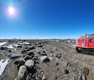 A red box-shaped vehicle towing a red box-shaped trailer, both on tank tracks, stopped on a dirt road in a rocky landscape