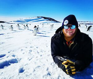 A smiling man wearing sunglasses and a beanie lies in the snow with about 20 penguins standing in the background