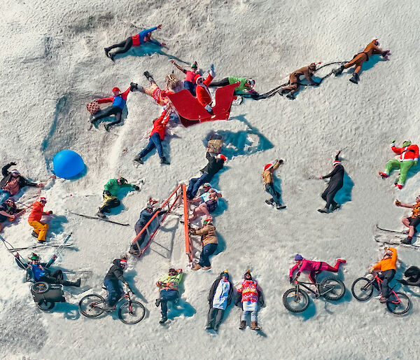 30 people, photographed from above, dressed in colourful Christmas costumes lying in the snow staging a Christmas scene with Santa, a sleigh, and elves