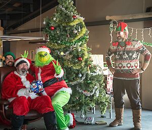 A man dressed as Santa gives a present to another man dressed as the Christmas Grinch. Two laughing men dressed as elves look on.